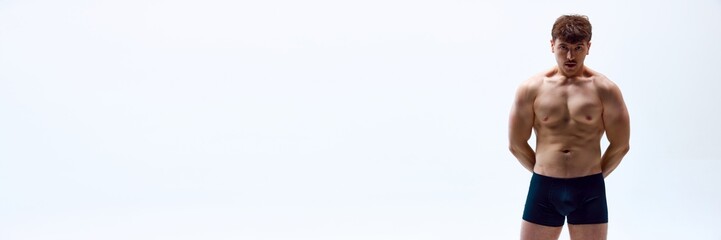 Fototapeta premium Portrait of handsome young man with muscular body posing shirtless in underwear on white studio background. Concept of man's beauty, sport and health, athletic body. Banner. Copy space for ad