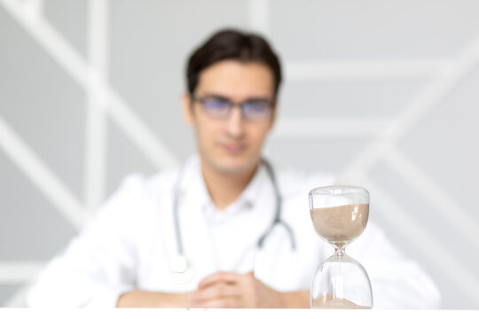 Close-up photo of hourglass on the desktop and male doctor on background. Time and health concept	