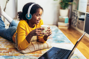 Smiling school African American girl in headphones watching the video lesson on computer,  happy child in earphones have online web class using laptop at home, homeschooling concept