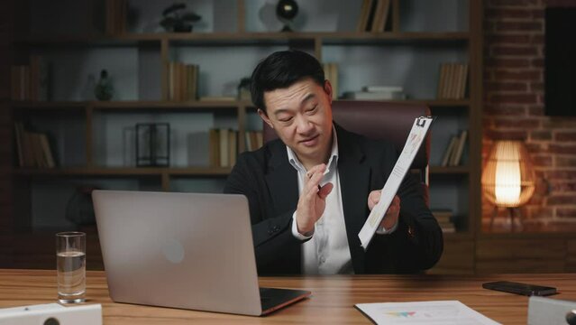 International manager taking part in online webinar with coworkers while sitting on chair in creative workplace. Diligent professional holding papers and pointing at issues that must be finalized.