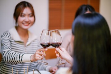 Cheerful friends enjoying home Birthday holiday party. Asian sister cheering drinking red wine celebrating with Birthday cake.