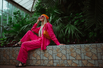 Fototapeta premium Fashionable happy smiling woman wearing trendy pink fuchsia color sunglasses, bandana, linen suit with oversized shirt, wide leg trousers, posing in tropical garden. Copy, empty space for text