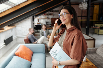 Young successful female manager or office worker with financial documents in hand talking on...