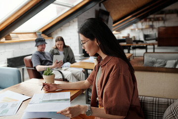 Side view of young brunette female economist in casualwear looking at paperwork with financial data while sitting by table in front of camera