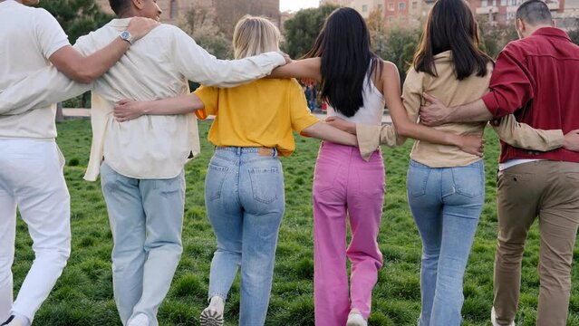 Multicultural group as team and community side by side in nature. Unrecognizable friends together in park hugging each other. Rear view photo of young people outdoors united in park. 