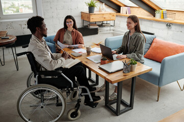 Two young intercultural female managers looking at confident African American male colleague in wheelchair during discussion at meeting