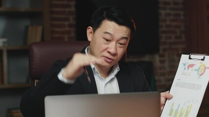 Strategic asian male executive presenting to coworkers papers with charts and graphs and making financial analysis. Proactive businessman conducting video conference using personal computer in office.