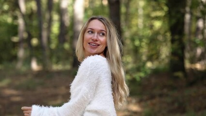 Portrait of relaxed happy woman looking at camera tossing long blond hair in slow motion standing in green forest. Beautiful Caucasian relaxed tourist with blue eyes posing in woodland on weekend