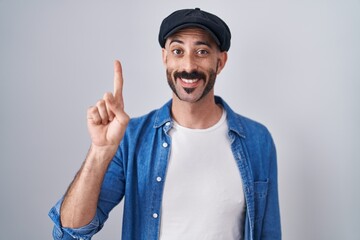 Hispanic man with beard standing over isolated background showing and pointing up with finger number one while smiling confident and happy.