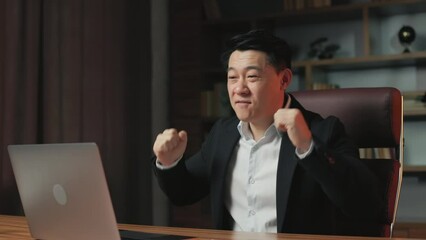 Ecstatic asian employee getting job promotion and celebrating work accomplishments in trendy office. Happy business man looking at laptop screen and dancing euphorically with hands on rolling chair.