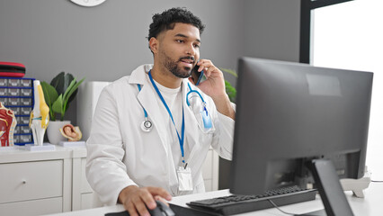 Fototapeta na wymiar African american man doctor using computer talking on smartphone at clinic
