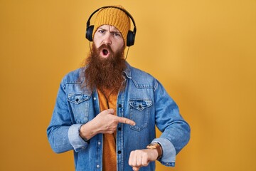 Caucasian man with long beard listening to music using headphones in hurry pointing to watch time,...