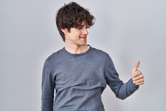 Young man standing over isolated background looking proud, smiling doing thumbs up gesture to the side