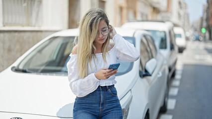 Young blonde woman using smartphone leaning on car at street