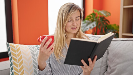 Young blonde woman reading book and drinking coffee sitting on sofa at home
