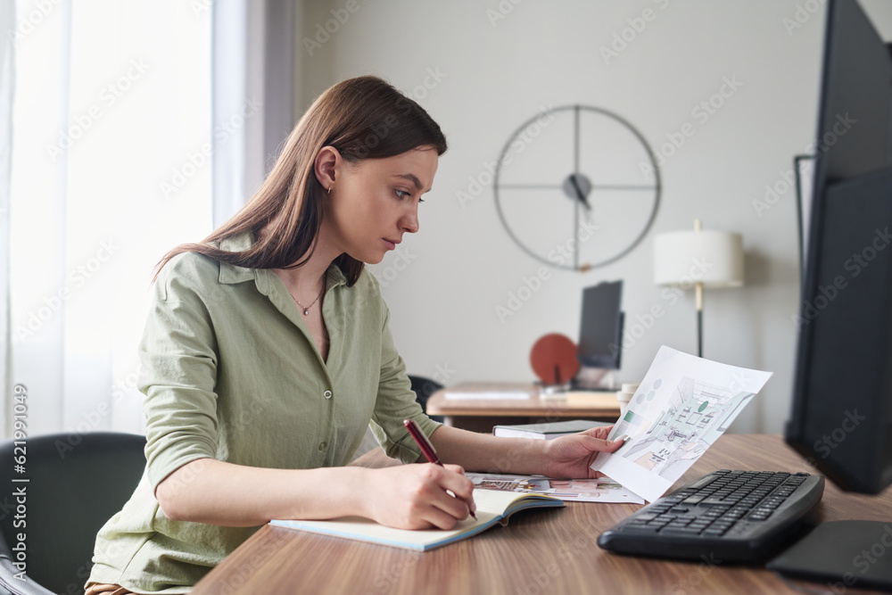 Aufkleber young serious female designer with pen over page of copybook looking at paper with sketch of interio