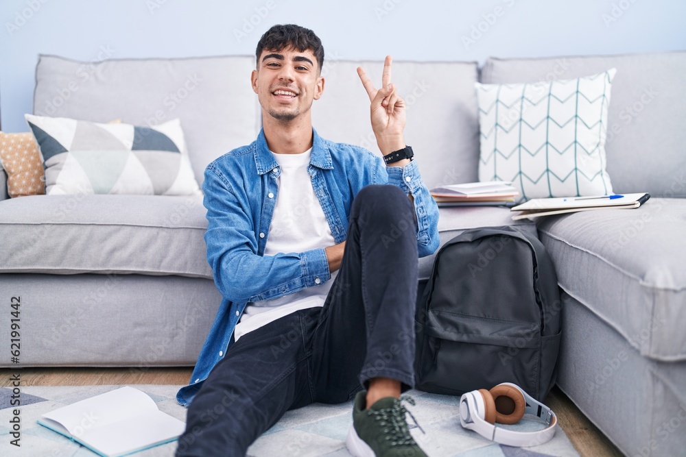 Wall mural Young hispanic man sitting on the floor studying for university smiling with happy face winking at the camera doing victory sign with fingers. number two.