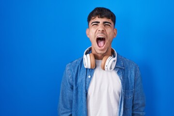 Young hispanic man standing over blue background angry and mad screaming frustrated and furious,...