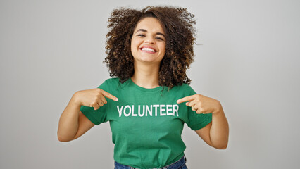 Young beautiful hispanic woman pointing to volunteer uniform smiling over isolated white background