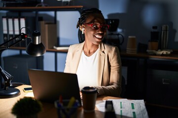 Beautiful black woman working at the office at night looking away to side with smile on face, natural expression. laughing confident.
