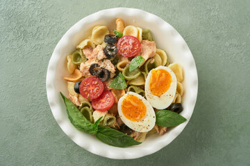 Italian pasta salad. Orecchiette pasta with tuna, tomato cherry, olive, basil and parmesan cheese in plate on green stone or concrete background. Traditional Italian cuisine. Flat lay. Copy space.