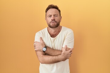 Middle age man with beard standing over yellow background shaking and freezing for winter cold with sad and shock expression on face