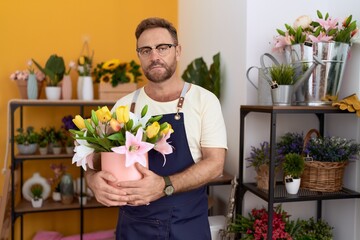 Middle age man with beard working at florist shop holding plant thinking attitude and sober expression looking self confident