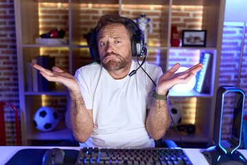Middle age man with beard playing video games wearing headphones clueless and confused expression...