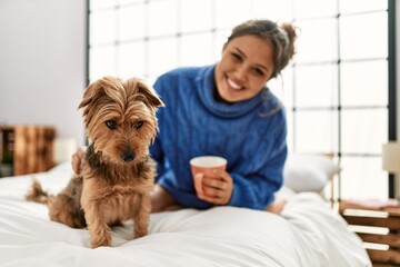 Young beautiful hispanic woman drinking cup of coffee sitting on bed with dog at bedroom