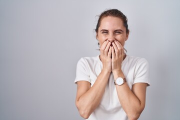 Beautiful brunette woman standing over isolated background laughing and embarrassed giggle covering...