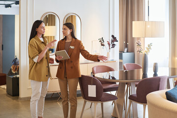 Young confident manager with tablet showing new pieces of furniture to female customer and helping...