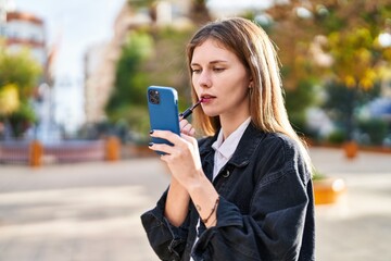 Young blonde woman using smartphone as a mirror make up lips at park