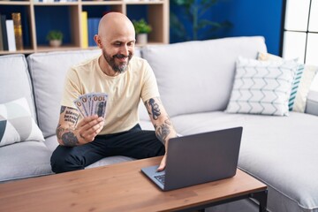 Young bald man using laptop holding dollars at home