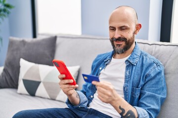 Young bald man using smartphone and credit card sitting on sofa at home