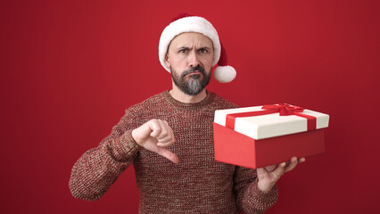 Young bald man wearing christmas hat holding gift looking upset over isolated red background