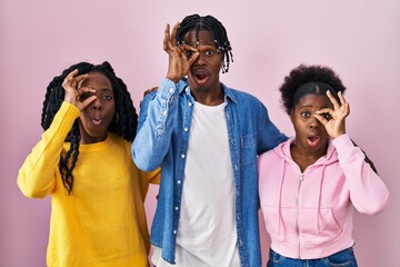 Group of three young black people standing together over pink background doing ok gesture shocked with surprised face, eye looking through fingers. unbelieving expression.