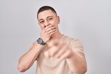 Young man standing over isolated background laughing at you, pointing finger to the camera with hand over mouth, shame expression