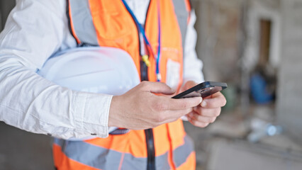 Young caucasian man architect using smartphone holding hardhat at construction site