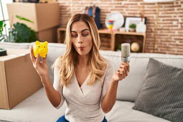 Young blonde woman holding led bulb and piggy bank making fish face with mouth and squinting eyes,...