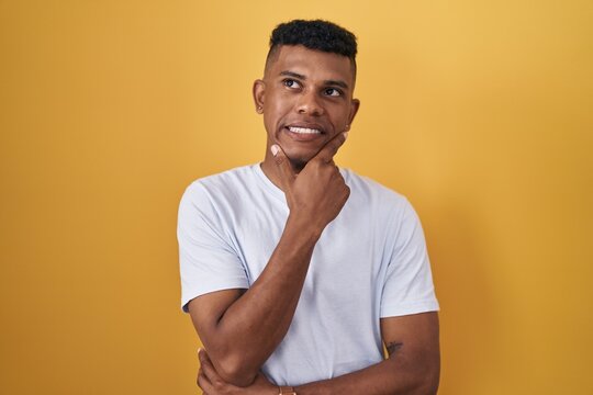 Young hispanic man standing over yellow background thinking worried about a question, concerned and nervous with hand on chin