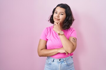 Young hispanic woman standing over pink background looking confident at the camera smiling with crossed arms and hand raised on chin. thinking positive.