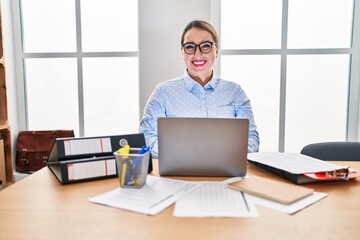 Fototapeta premium Young hispanic woman working at the office wearing glasses looking positive and happy standing and smiling with a confident smile showing teeth