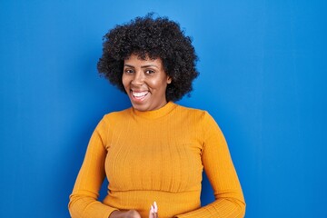 Fototapeta na wymiar Black woman with curly hair standing over blue background with hands together and crossed fingers smiling relaxed and cheerful. success and optimistic