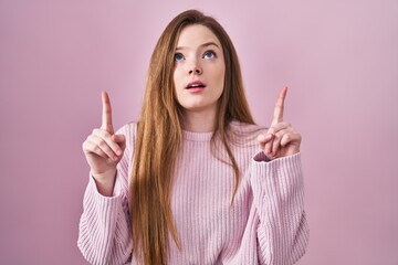 Young caucasian woman standing over pink background amazed and surprised looking up and pointing with fingers and raised arms.