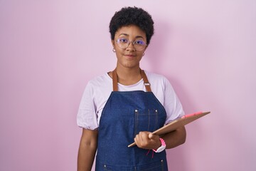 Young african american woman wearing professional waitress apron holding clipboard relaxed with...