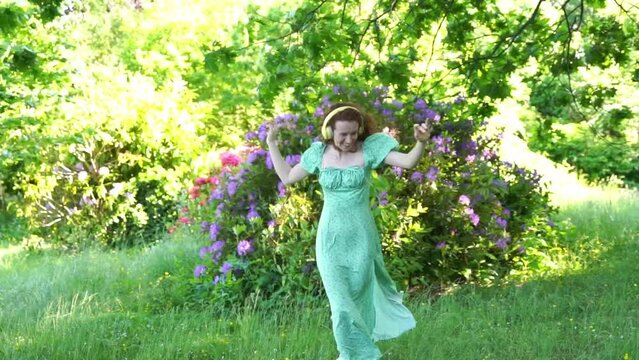 Outdoor portrait of happy red hair woman in green dress and headphones dancing in the public park. Listen to online music anywhere and be yourself concept	