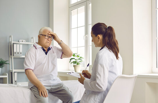 Senior man patient suffering from a headache, sitting on a medical couch, holding hand on his head and telling the doctor about the intense pain that he is feeling. Medicine, medical help concept