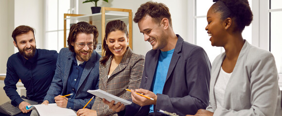 Business team having fun at a work meeting. Diverse group of happy cheerful young people in smart casual clothes sitting in the office, brainstorming, writing down ideas and smiling. Banner background © Studio Romantic