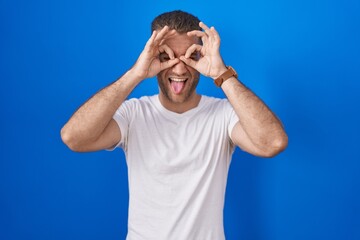 Young caucasian man standing over blue background doing ok gesture like binoculars sticking tongue out, eyes looking through fingers. crazy expression.