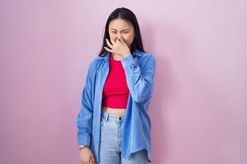 Young asian woman standing over pink background smelling something stinky and disgusting, intolerable smell, holding breath with fingers on nose. bad smell